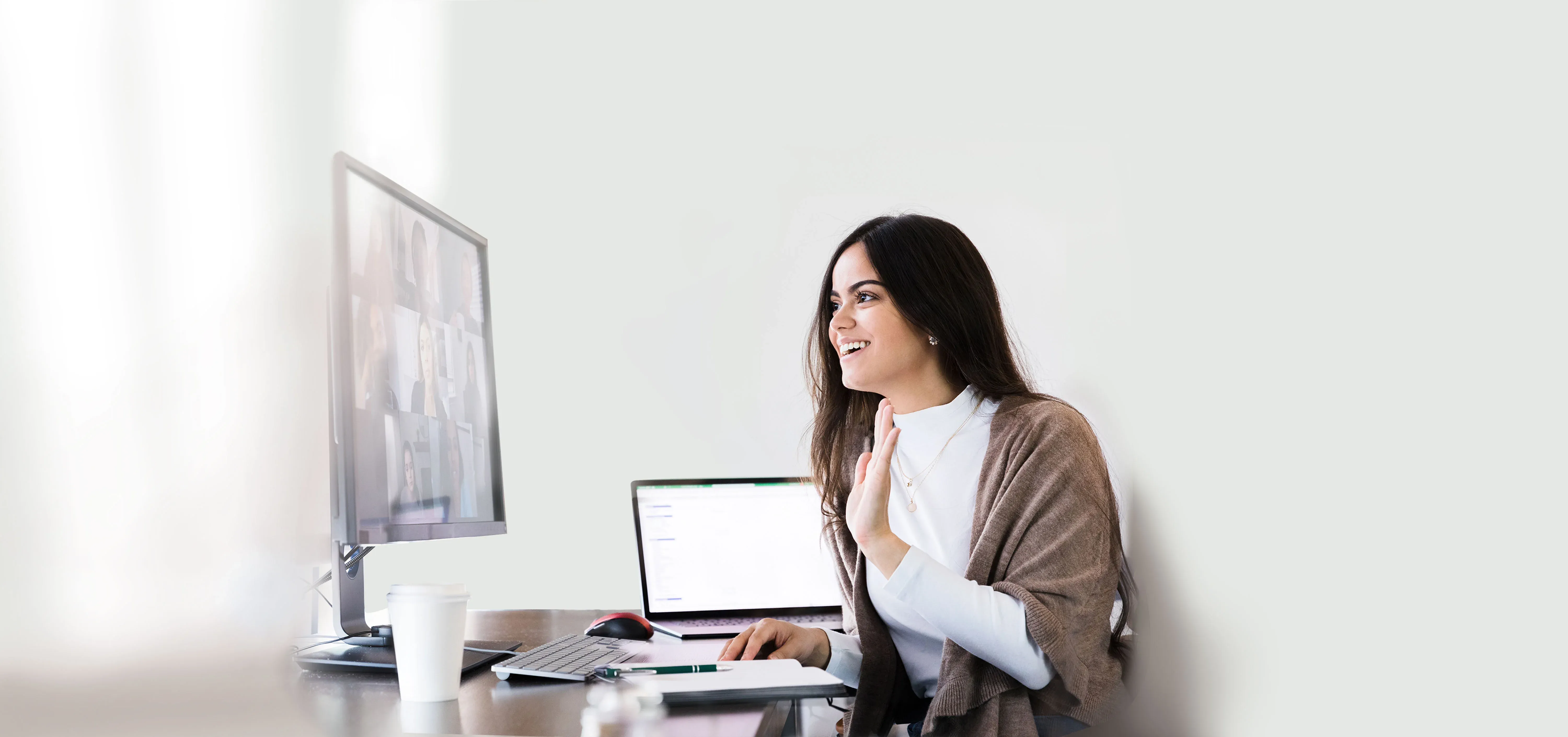 Young professional woman waving and smiling while engaged in a video conference call where we can see the screen (although it is blurred).